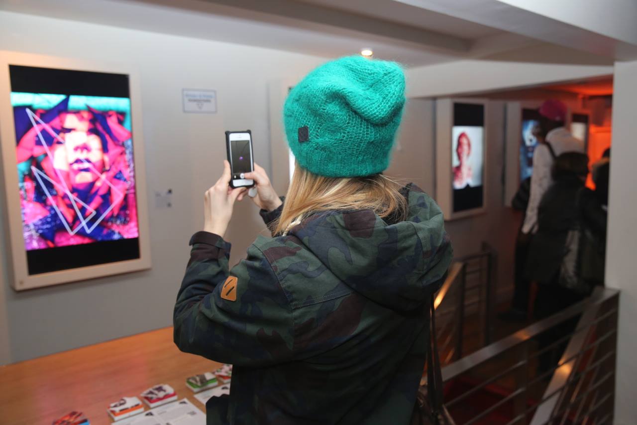 A person wearing a green beanie and camo jacket is snapping a photo with a smartphone at an Arthouse gallery exhibition in SoHo. The room is softly lit, showcasing digital artwork on the wall with two more pieces visible in the background, as other visitors admire the creative display.