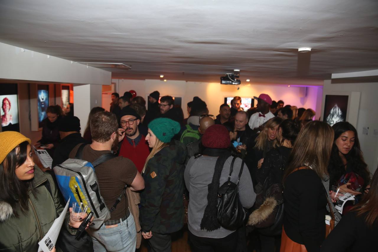 A crowded SoHo gallery exhibition features people mingling and engaging with displayed art on the walls. Artworks, illuminated by spotlights, add color to the dimly lit room. Attendees, dressed in winter clothing such as coats and hats, are conversing and holding brochures, creating a lively atmosphere.