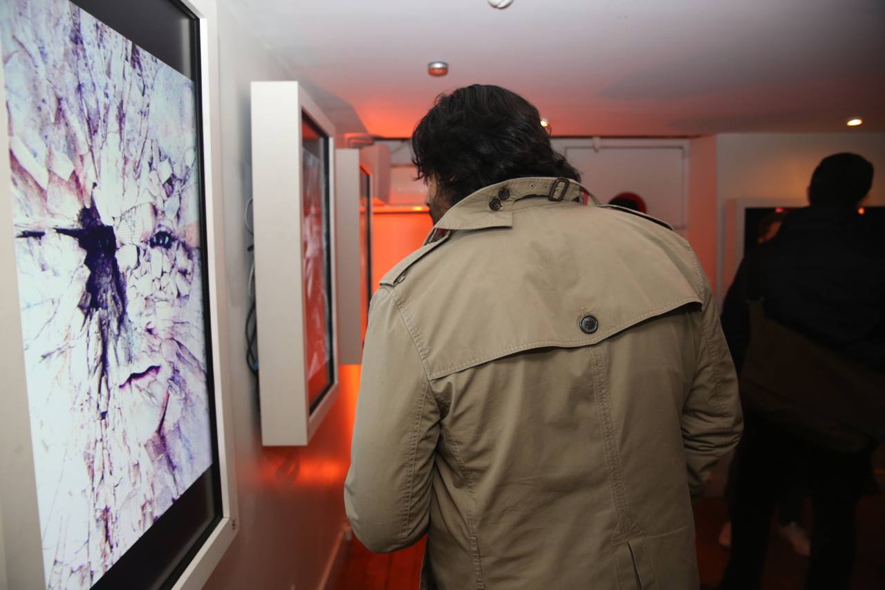 A person in a beige trench coat views a digital art display at the Arthouse gallery exhibition in SoHo. The artwork features abstract, cracked patterns with purple hues. Other visitors linger in the background as the dimly lit room is warmed by soft lighting.
