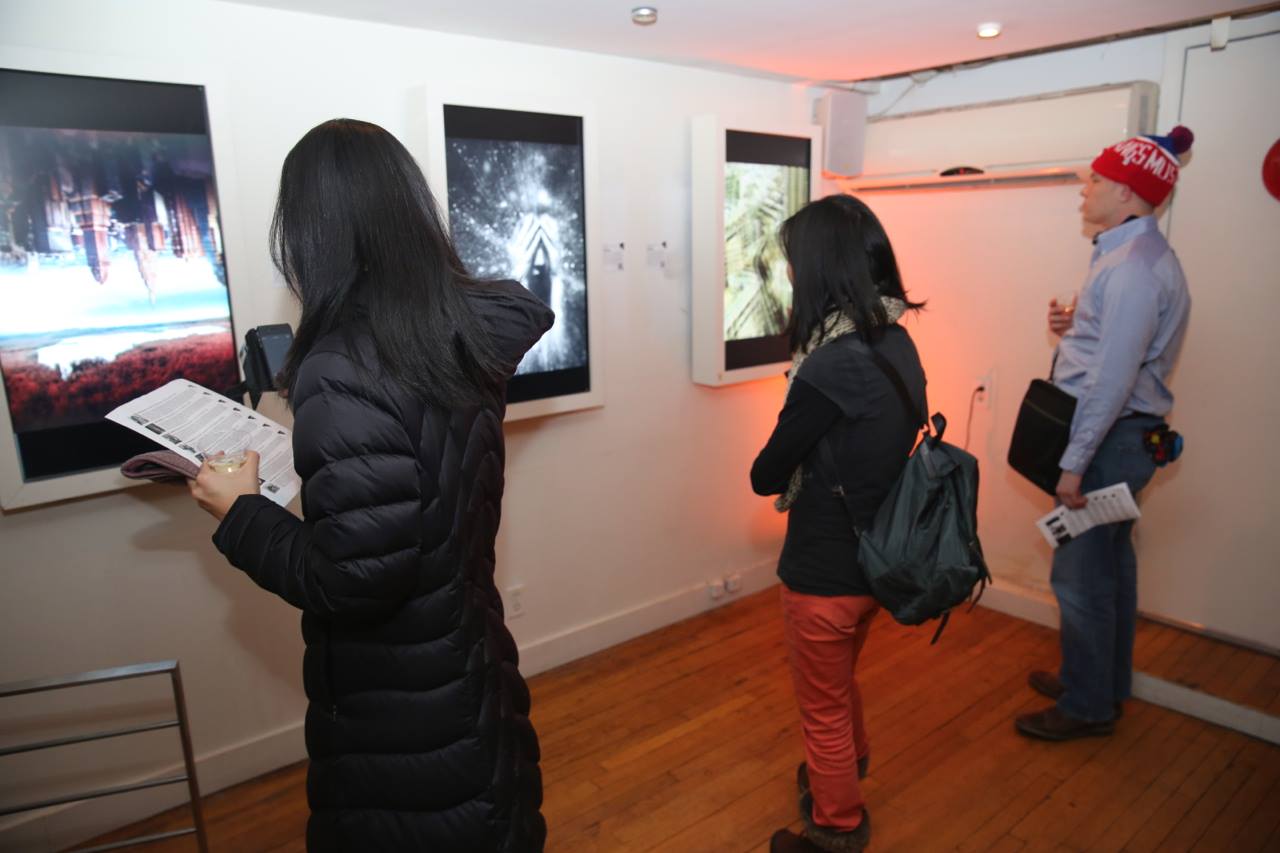 Three people explore an arthouse in SoHo. Two women examine digital displays; one holds a brochure, the other snaps a photo. A man in a red beanie observes another piece. Hardwood floors and white walls create a modern, cozy atmosphere at this gallery exhibition.