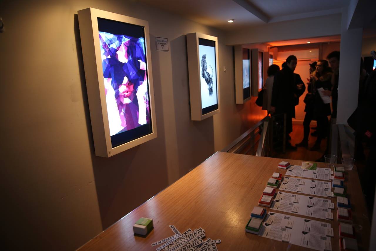 A dimly lit SoHo art gallery room features digital art displays on the wall. A wooden table in the foreground holds papers and small stacks of colored cards, with name tags scattered on it. In the background, people are gathered, engaged in conversation near softly illuminated walls.