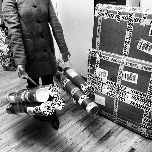 A person in a coat carries several rolls of wrapping paper in one hand while standing on a wooden floor. Next to them are three large, sealed cardboard boxes labeled 