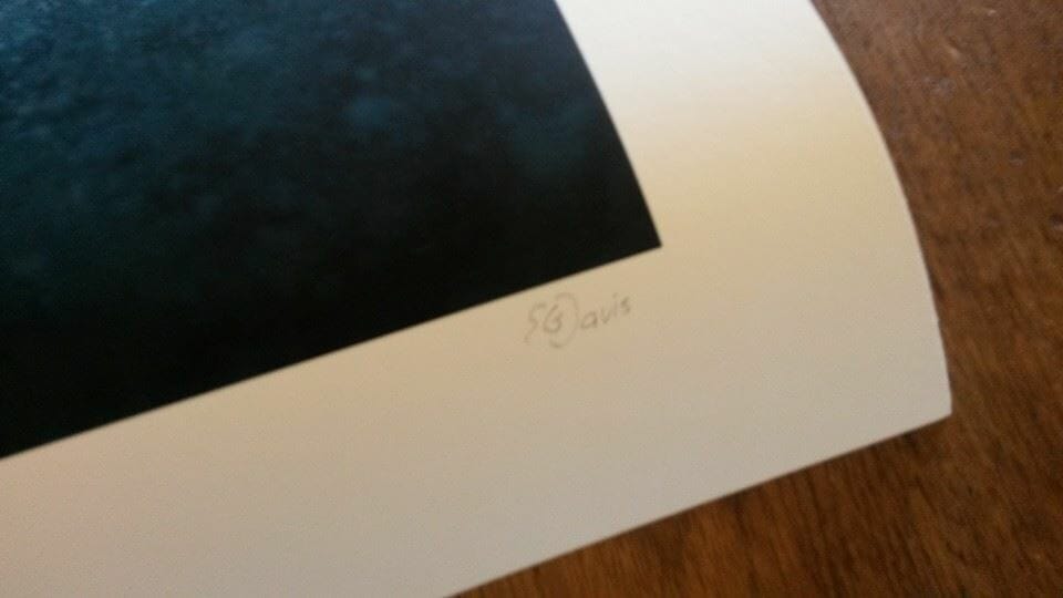 Close-up of the lower right corner of a photograph on a wooden table. The edge is white, and the dark image is mostly out of view. 