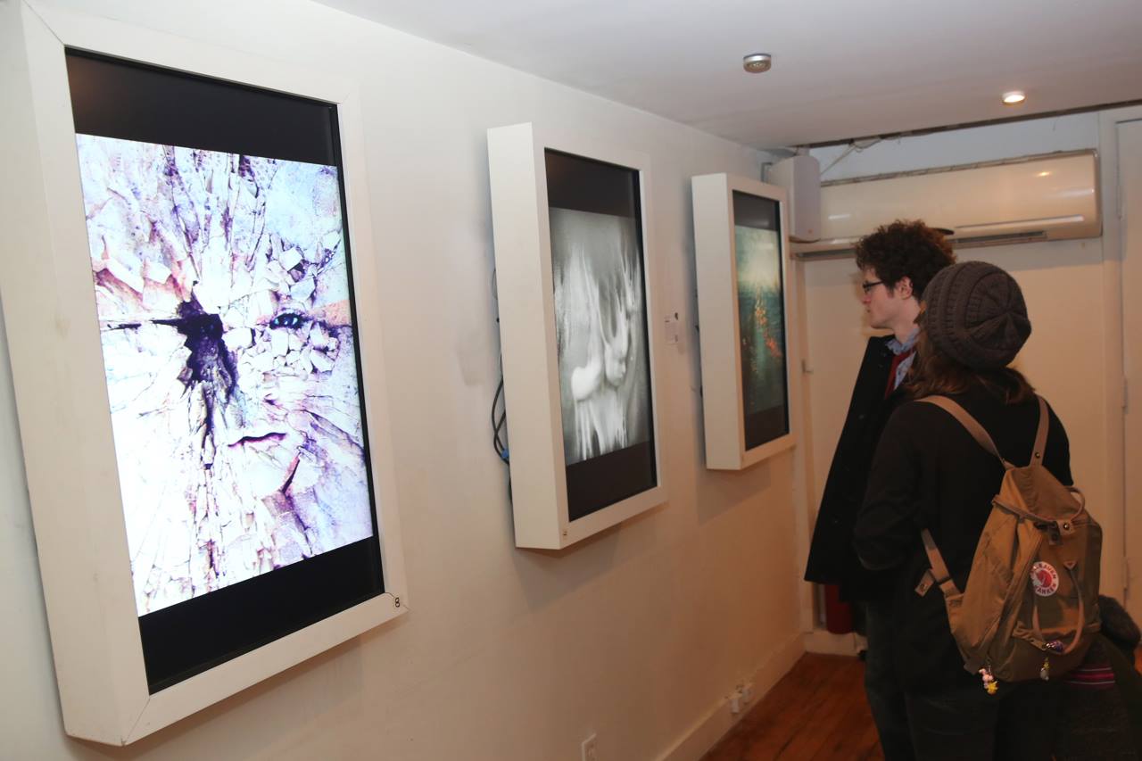 Two people, one wearing a beanie hat and backpack, and the other with curly hair and glasses, stand in an Arthouse gallery exhibition in SoHo, observing digital artworks displayed on large vertical screens mounted on the wall. The artworks feature abstract designs and are brightly illuminated.