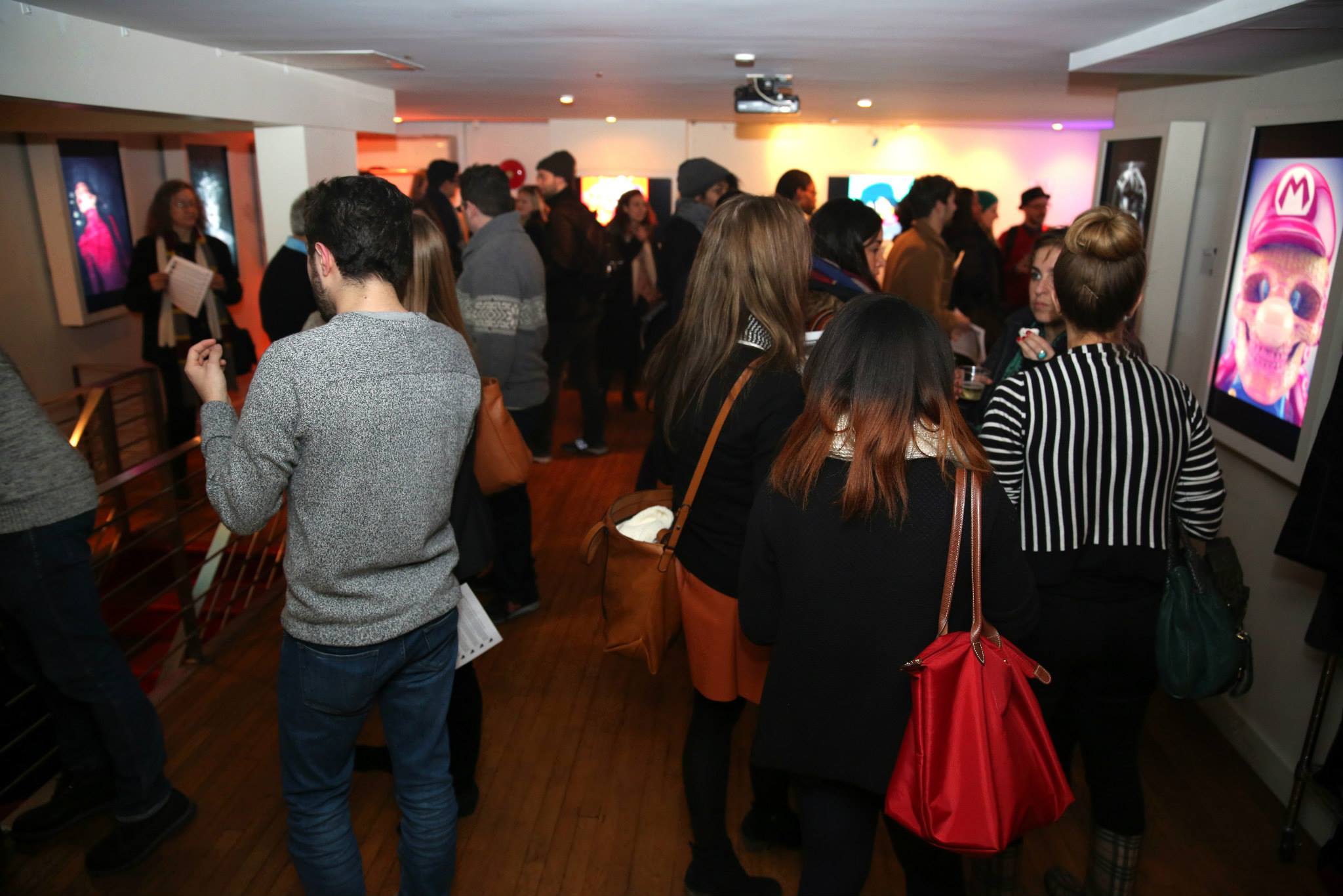 A group of people mingling at an indoor social event in a SoHo arthouse gallery. Attendees are casually dressed and engaged in conversation, some holding drinks. The room is dimly lit with spotlights on several framed images on the walls. There are wooden floors, white walls, and a projection screen in the background.
