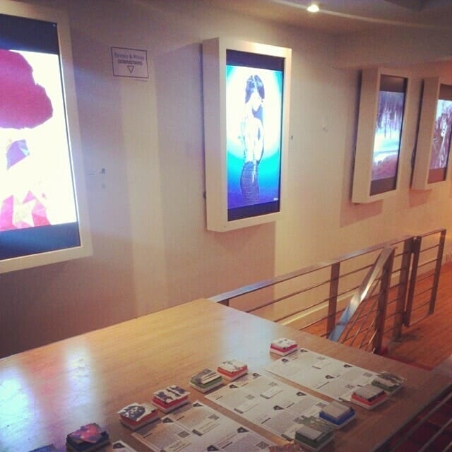 The SoHo Arthouse presents a modern gallery space with four illuminated digital displays showcasing colorful abstract and photographic art. Below, a wooden table holds neatly arranged stacks of brochures for the gallery exhibition. The space features wooden flooring and metal railings.